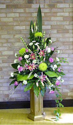 a floral arrangement in a gold vase on the floor next to a brick wall,