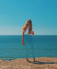 a woman standing on top of a sandy beach next to the ocean with her hair blowing in the wind