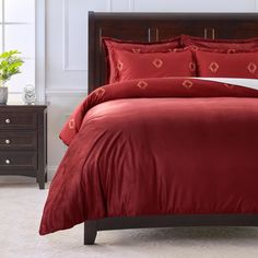 a bed with red comforter and pillows in a room next to a dresser, potted plant and window