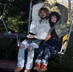 two people are sitting on a swing in the park, one is holding his arm around the other's shoulder