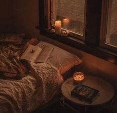 an open book on a bed next to two candles and a table in front of a window