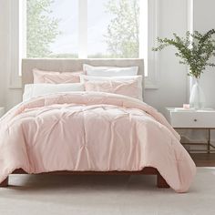 a bed with pink comforter and pillows in front of a large window, next to a plant