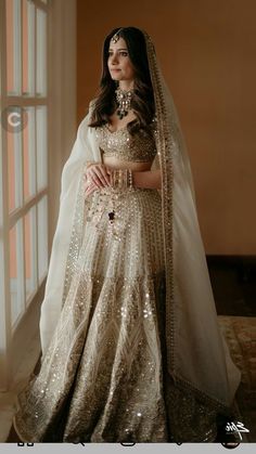 a woman in a white and gold bridal gown standing by a window with her hands on her hips