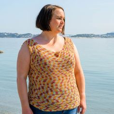 a woman standing by the water with her hands in her pockets, wearing a yellow and red crochet top