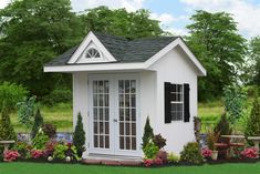 there is a small white shed in the grass with flowers around it and trees behind it