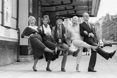 an old black and white photo of five people posing on the sidewalk with their legs in the air