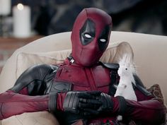 a deadpool sitting in a chair with his arm around a white cat that is looking at him