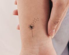 a small dandelion tattoo on the wrist is being held by a woman's hand