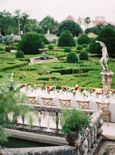 an outdoor dining area with tables and chairs in front of a formal garden setting, surrounded by greenery