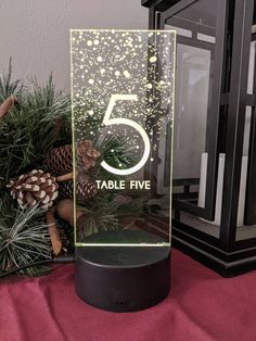 an illuminated table number with pine cones and evergreens in front of a christmas tree