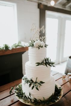 a white wedding cake sitting on top of a wooden table with greenery around it