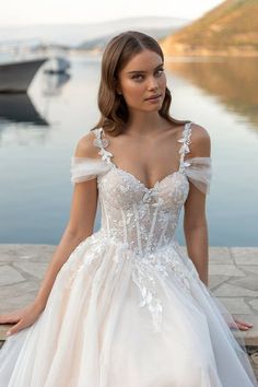 a woman in a wedding dress is sitting on the ground near some water and boats