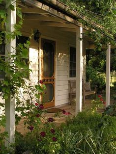 the front porch of a small white house surrounded by greenery and flowers on a sunny day