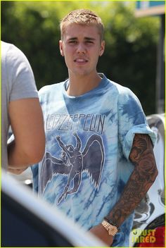 a young man with tattoos on his arm is standing in front of a car and looking at the camera