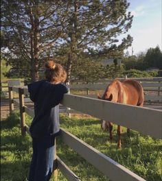 a young boy standing in front of a fence looking at a horse grazing on the grass