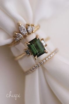 two rings on top of each other with green stones and diamonds around them, sitting on a white cloth