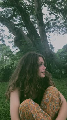 a woman with long hair sitting on the ground in front of a tree and grass