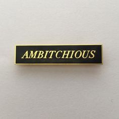 a black and gold pin with the words ambitioushouse on it's back side