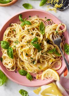 a pink plate topped with pasta and lemon wedges next to a bowl of basil