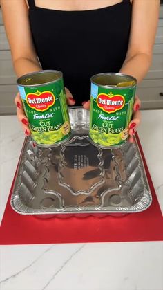 a woman holding two cans of canned food on top of a metal pan with red mat