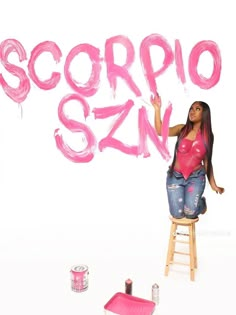 a woman is sitting on a stool and painting the word scorpio san in pink