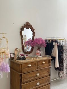 a dresser and mirror in a room with clothes hanging on the rack, and flowers