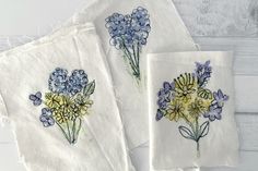 three embroidered napkins with blue and yellow flowers on them, sitting on a white surface