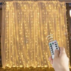 a hand holding a remote control in front of a curtain with lights on the outside