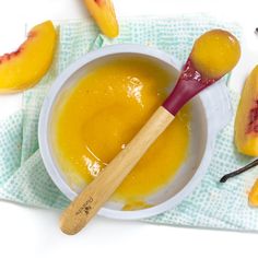 a white bowl filled with yellow liquid next to sliced peaches and a wooden spoon