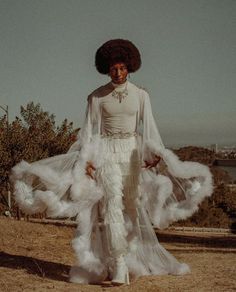 a woman with an afro standing on top of a hill wearing a white dress and feathers