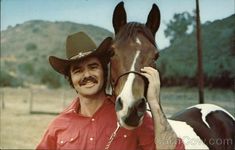 a man in a cowboy hat standing next to a brown and white horse wearing a red shirt