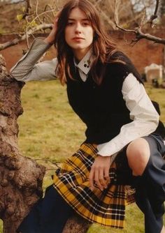 a young woman sitting on top of a tree branch wearing a plaid skirt and sweater