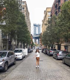 a woman walking down a street next to parked cars and tall buildings with a bridge in the background