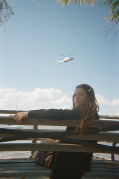 a woman sitting on top of a wooden bench next to the ocean with a plane flying overhead