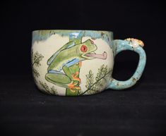 a ceramic mug with a frog painted on it's front and side, sitting on a black surface