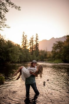 a man holding a woman in the water near some trees and mountains at sunset or dawn