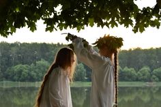 two women dressed in white standing next to each other on a tree swing by the water