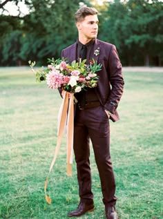 a man in a suit and tie holding a bouquet of flowers on the grass with trees in the background