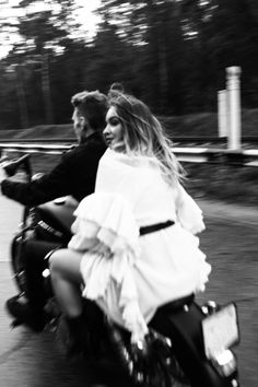 a man and woman riding on the back of a motorcycle down a road in black and white