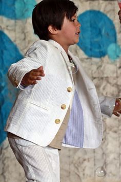 a young boy in a white suit and tie holding a wine glass while standing next to a wall