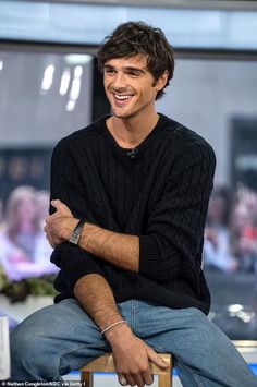 a smiling man sitting on top of a wooden chair in front of a tv screen