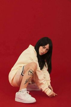 a woman with black hair and tattoos on her leg, crouching down in front of a red background