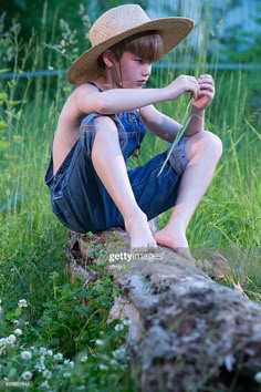 a young boy sitting on top of a log in the grass wearing a straw hat