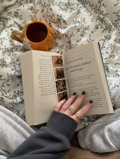 a woman laying in bed reading a book and holding a coffee mug with both hands