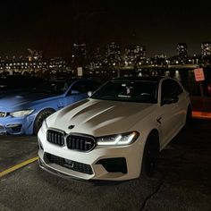 two bmws parked in a parking lot at night with city lights behind them and buildings in the background