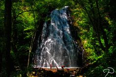 two people standing at the base of a waterfall surrounded by green foliage and trees with water cascading over it