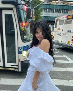 a woman in a white dress is crossing the street with a bus behind her on a city street