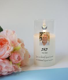 a white candle with a photo on it next to pink flowers and a blue background