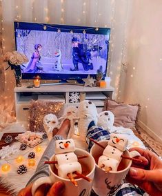 two people are holding mugs with marshmallow faces on them in front of a television