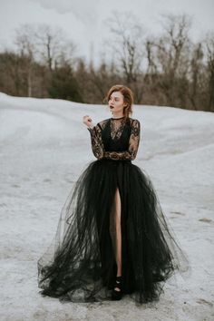 a woman standing in the snow wearing a black dress with a high slit and long sleeves